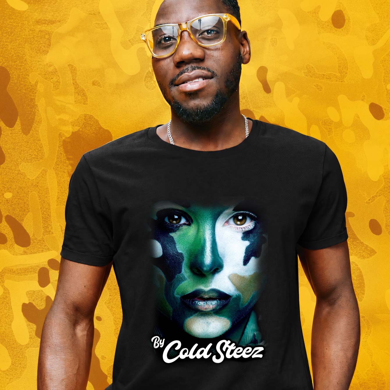 A man wearing a black Streetwear t-shirt with a stylized graphic of a woman's face in green, blue, and white, camo with the text 'By Cold Steez' printed below. The background is a textured yellow and gold abstract pattern.